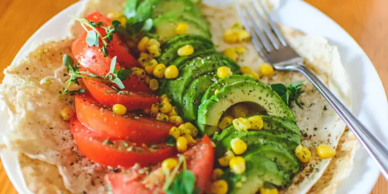 Plate of avocado toast topped with sliced avocado, tomato, and corn, garnished with herbs on a white plate