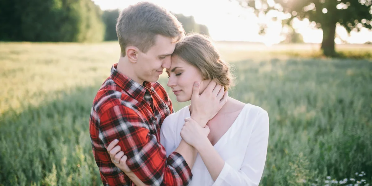 Couple embracing in a sunlit field. The man in a red plaid shirt cradles the woman's neck as they lean their foreheads together.