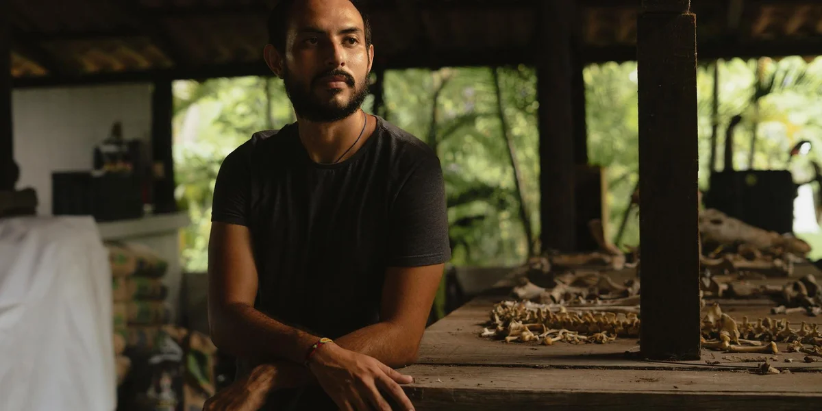 A man stands in a dim room beside a long table with reptile enclosures and care supplies, illustrating a home gecko care setup.