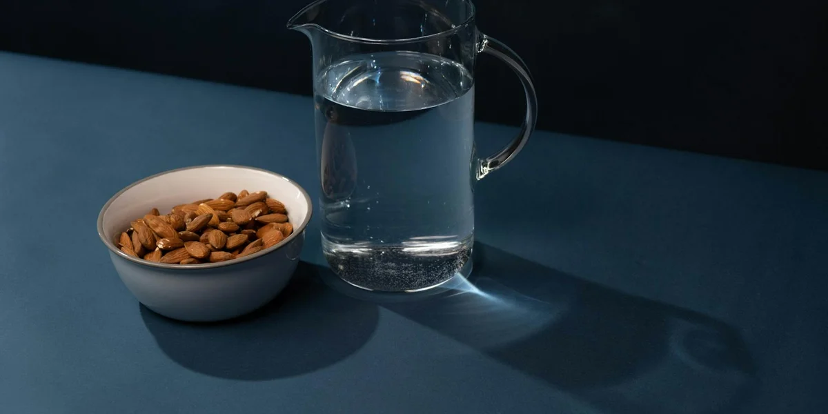 A clear glass pitcher of water beside a small white bowl of almonds on a blue tabletop, with soft shadows.