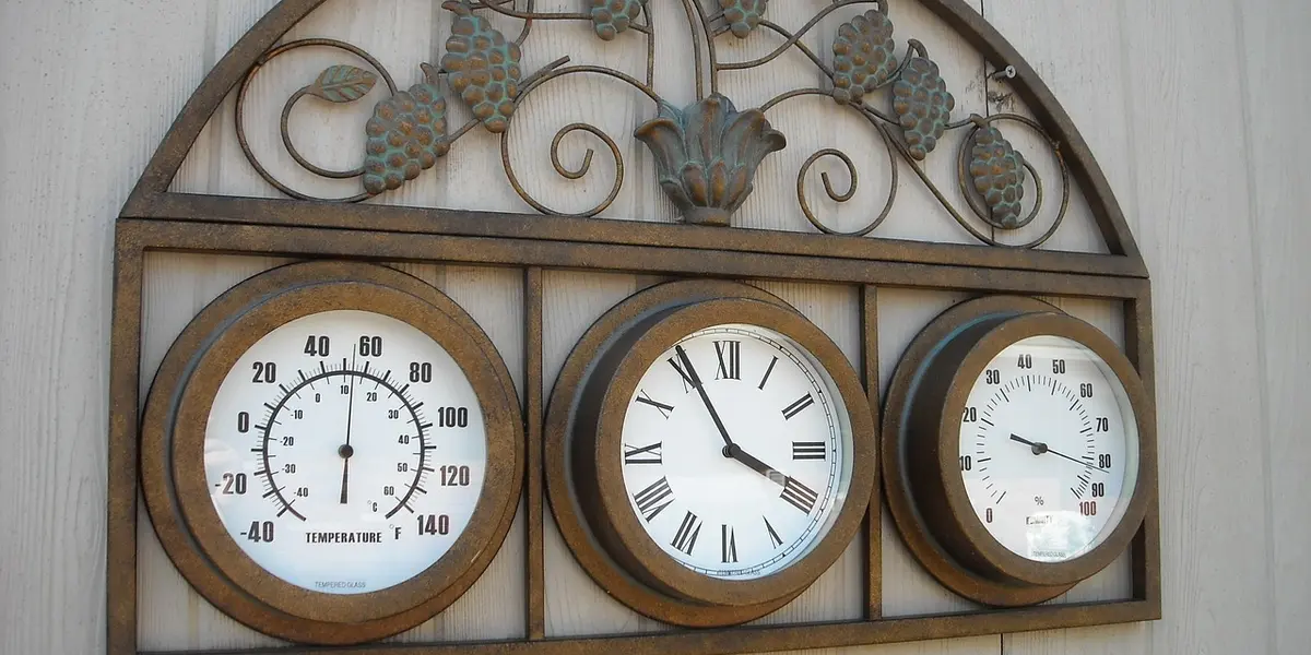 Three round gauges mounted in a decorative rustic metal frame: a thermometer on the left, a clock in the center, and a third gauge on the right.