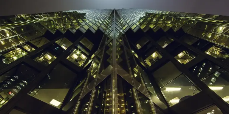 Nighttime view of a tall glass office building with mirrored windows forming a geometric pattern, reflecting city lights.