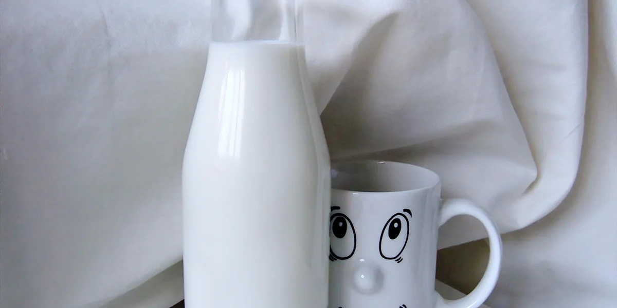 White milk bottle beside a white mug with a cartoon face on a soft white fabric background.