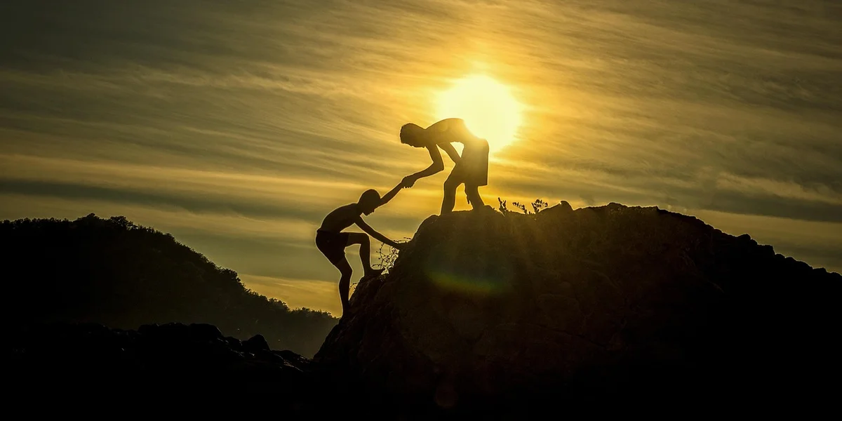 Two silhouetted figures helping each other climb a rocky ridge at sunset.