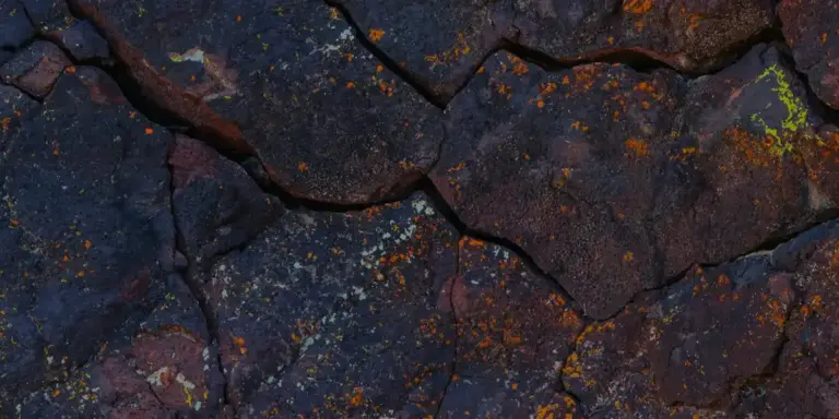 Close-up of a dark, textured rock surface with orange-brown rust-like patches and small greenish lichen.