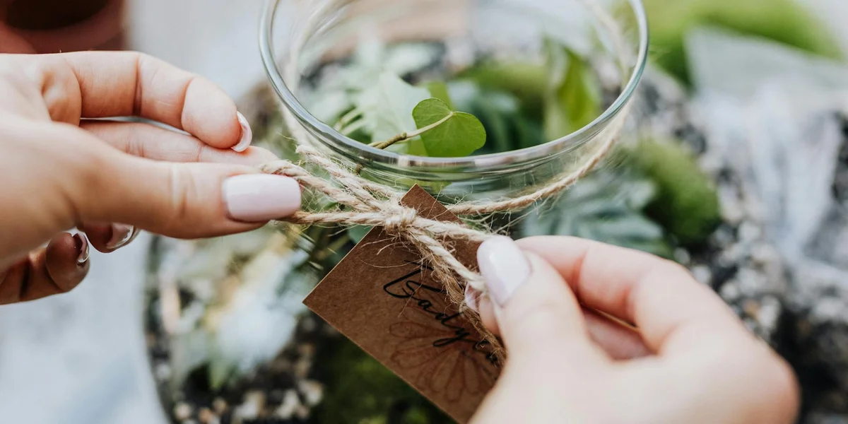 Close-up of hands tying a twine tag to a glass jar filled with fresh plant cuttings, used as decor in a terrarium.