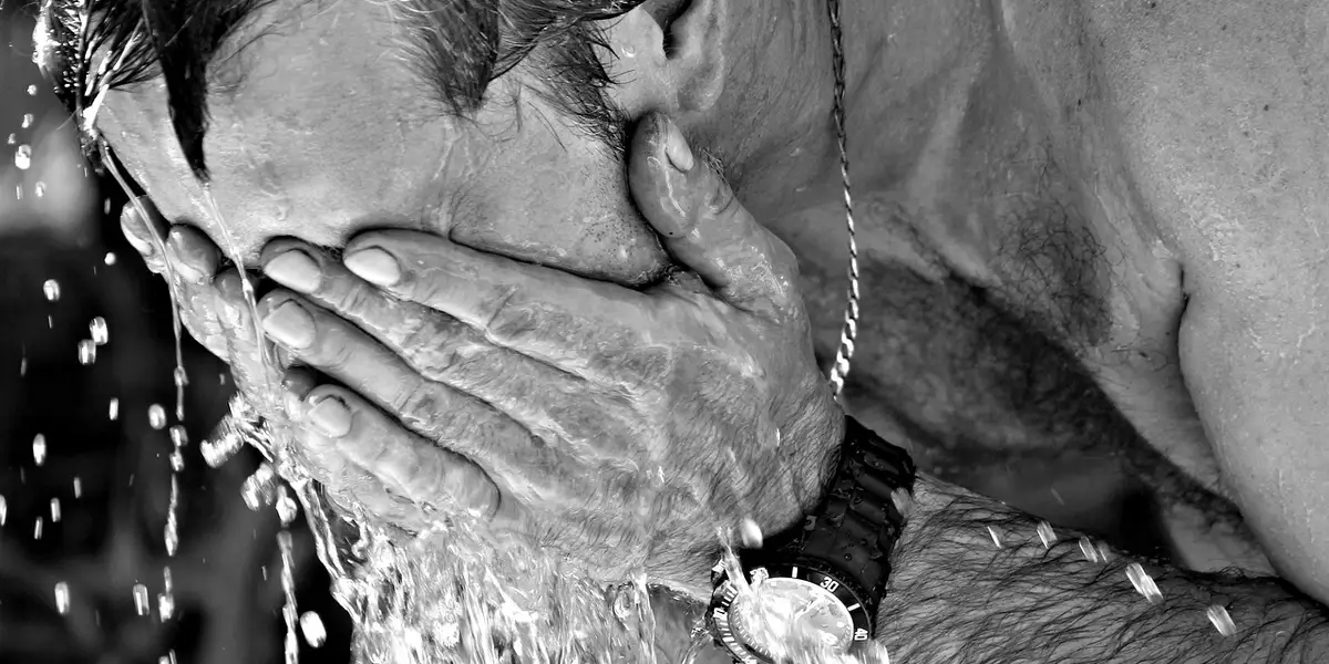 Close-up of a person covering their face with wet hands under spraying water, illustrating thermoregulation stress.