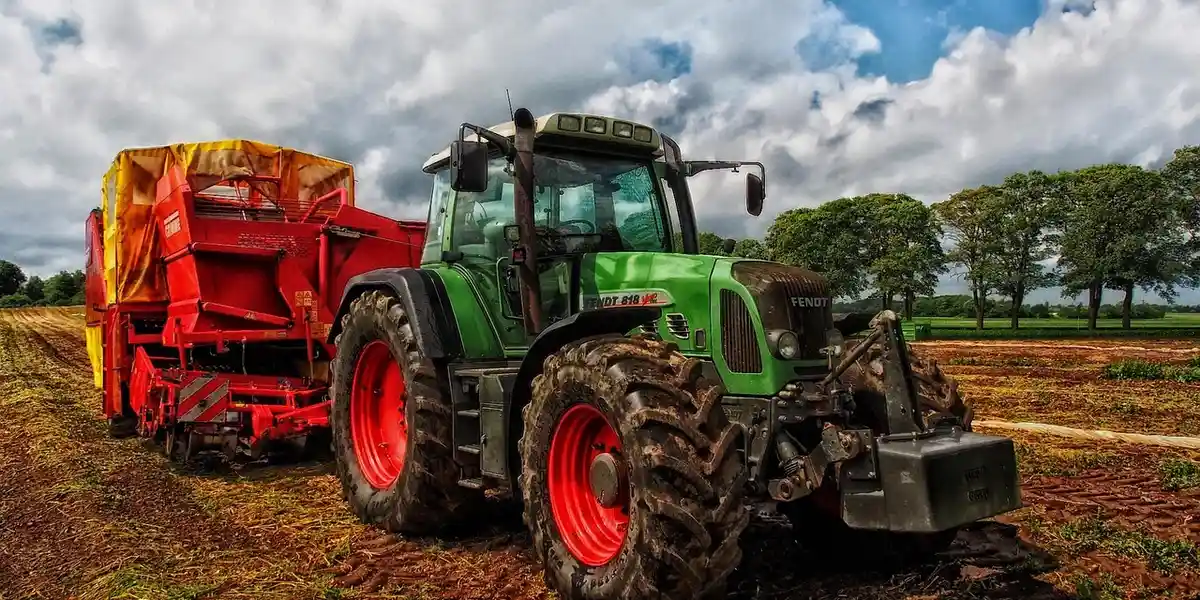 Green tractor pulling a red harvesting trailer on a plowed field with trees in the background under a cloudy sky.